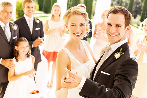 Portrait of happy newlywed couple dancing with wedding guests applauding in background during reception. Horizontal shot.